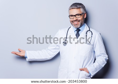 Doctor advertising your product. Cheerful mature doctor looking at camera and pointing away while standing against grey background