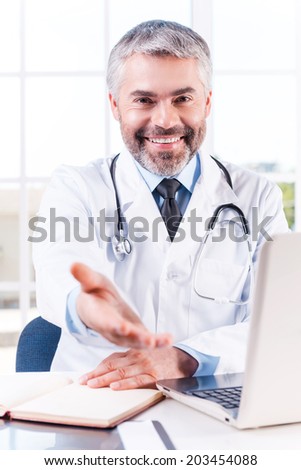 You can trust me! Confident mature grey hair doctor stretching out hand and smiling while sitting at his working place