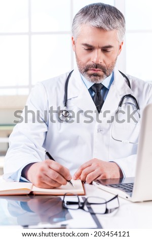 Confident doctor at work. Mature grey hair doctor writing something in note pad while sitting at his working place