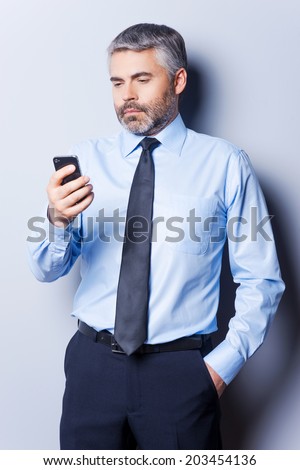 Checking out business messages. Confident mature man in shirt and tie holding mobile phone and looking at it while standing against grey background