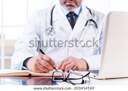 Confident doctor at work. Close-up of mature doctor writing something in note pad while sitting at his working place