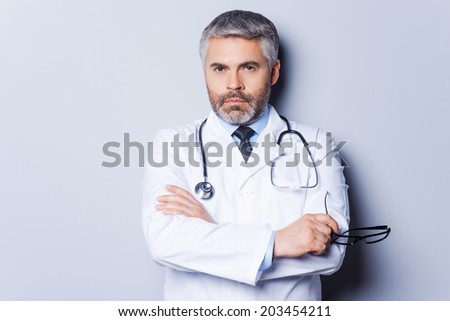 Confident doctor. Mature grey hair doctor looking at camera and keeping arms crossed while standing against grey background
