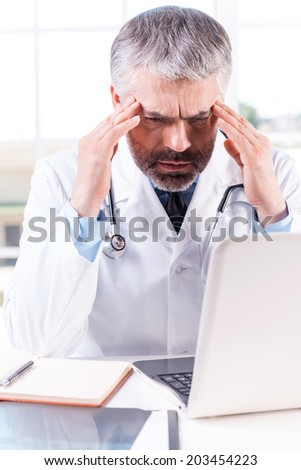 Stressed and tired doctor. Depressed mature grey hair doctor touching his head with hands while sitting at his working place
