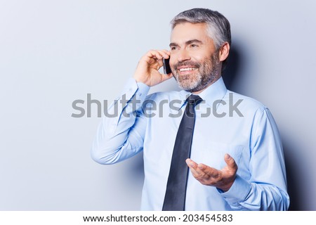 Sharing good news. Happy mature man in shirt and tie talking on the mobile phone and gesturing while standing against grey background