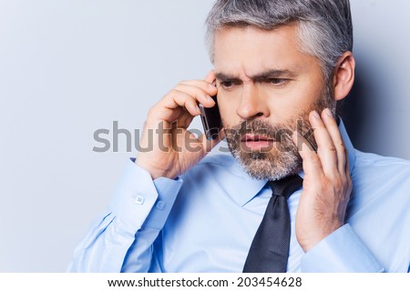 Bad news. Depressed mature man in shirt and tie talking on the mobile phone and touching face with hand while standing against grey background