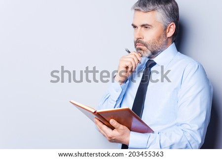 Waiting for inspiration. Thoughtful mature man in shirt and tie touching his chin with hand and looking away while holding note pad and standing against grey background