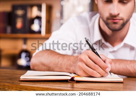 Confident bar owner. Close-up of handsome young male bartender in white shirt leaning at the bar counter and writing something in note pad