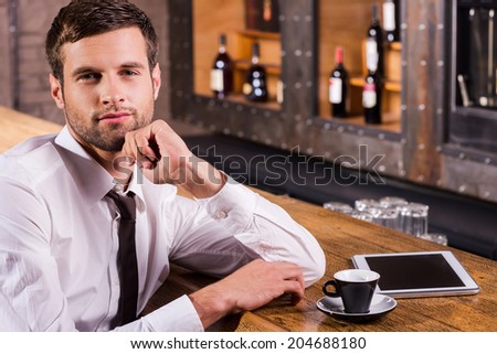 Surfing the net in bar. Handsome young man in shirt and tie holding hand on chin and smiling while sitting at the bar counter with digital tablet laying near him