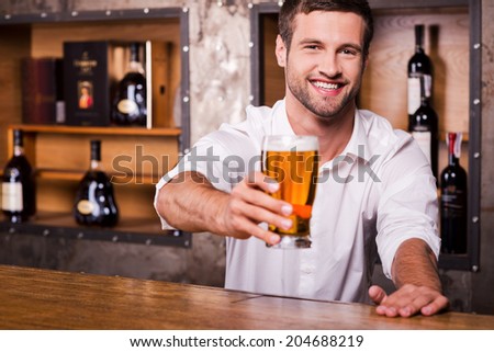 Let me quench your thirst! Happy young male bartender in white shirt stretching out glass with beer and smiling while standing at the bar counter
