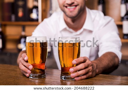 Quench your thirst! Close-up of handsome young male bartender in white shirt stretching out glasses with beer and smiling while standing at the bar counter