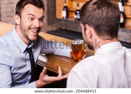 Sharing beer with good friend. Two cheerful young men in shirt and tie talking to each other and gesturing while drinking beer at the bar counter