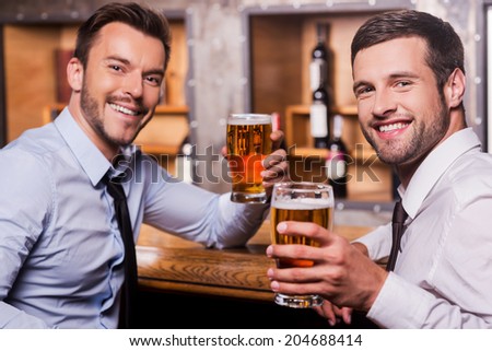 Relaxing after hard working day. Two happy young men in shirt and tie holding glasses with beer and smiling while sitting at the bar counter