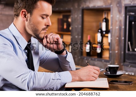 Writing down his ideas. Side view of handsome young man in shirt and tie writing something in note pad while sitting at the bar counter