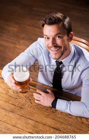 Celebrating success. Top view of handsome young man in shirt and tie holding glass with beer and smiling while sitting at the bar counter
