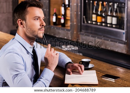 Waiting for inspiration. Side view of thoughtful young man in shirt and tie touching his chin with pen and looking away while sitting at the bar counter with note pad laying on it