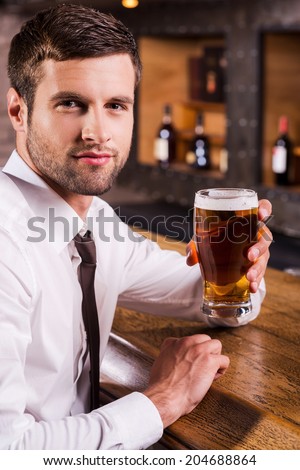 Refreshing with glass of cold beer. Side view of handsome young man in shirt and tie holding glass with beer and looking at camera while sitting at the bar counter