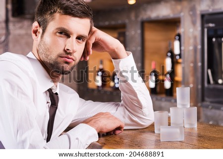 Man in bar. Handsome young man in shirt and tie leaning at the bar counter and and looking at camera while empty glasses standing near him
