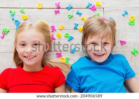 Cheeky kids. Top view of two cute little children looking at camera and smiling while lying on the floor with plastic colorful letters laying around them