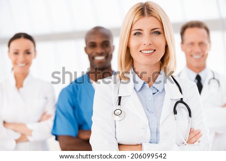 Confident in her team. Beautiful female doctor keeping arms crossed and smiling while her colleagues standing behind her in the background
