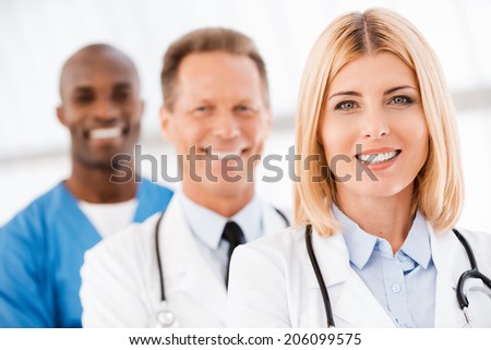 Medical doctors team. Confident female doctor looking at camera and smiling while her colleagues standing in a row behind her