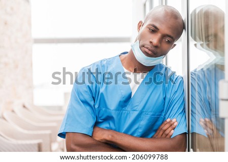 Depressed surgeon. Depressed young African doctor in blue uniform keeping arms crossed and looking away while leaning at the glass door