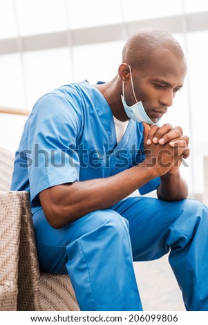Worried surgeon. Thoughtful young African doctor in blue uniform leaning his face on hands and looking down while sitting at the chair in corridor