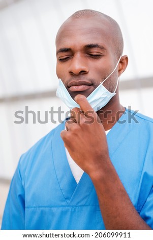 Tired after long surgery. Portrait of tired young African doctor in blue uniform taking of his surgical mask and keeping eyes closed
