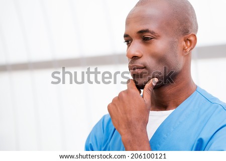 Confident and concentrated. Portrait of thoughtful young African doctor in blue uniform holding hand on chin and looking away