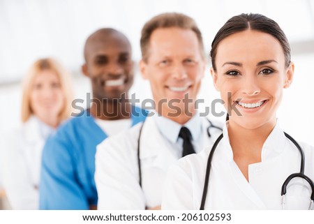 Confident doctors team. Confident female doctor looking at camera and smiling while her colleagues standing in a row behind her
