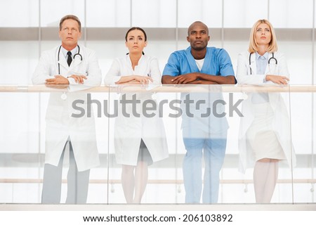 Confident team of medical experts. Low angle view of four confident doctors standing close to each other and looking away while leaning at the handrail