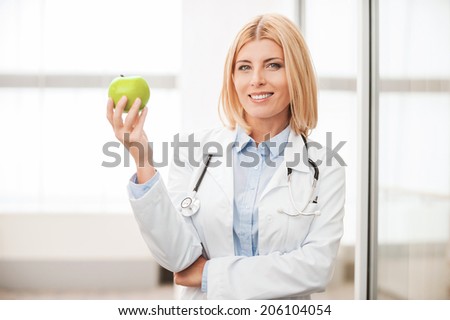 Eat healthy! Confident female doctor in white uniform holding green apple and smiling while leaning at the glass wall