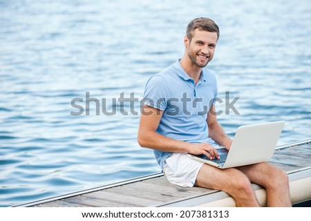 I like working outdoors! Handsome young man in polo shirt working on laptop and smiling while sitting on quayside