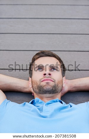 Man daydreaming. Top view of thoughtful young man holding hands behind head and looking away while lying down on the hardwood floor