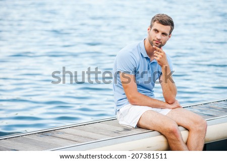 Handsome day dreamer. Thoughtful young man holding hand on chin and looking away while sitting at the riverbank