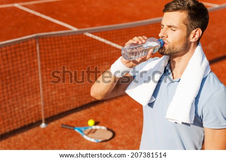 Getting refreshed after game. Thirsty young man in polo shirt and towel on shoulders drinking water while standing on tennis court
