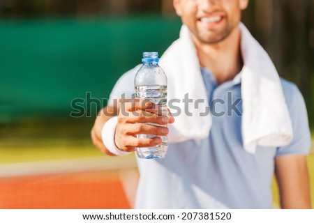 Drink some water! Close-up of happy young man in polo shirt and towel on shoulders stretching out bottle with water while standing on tennis court