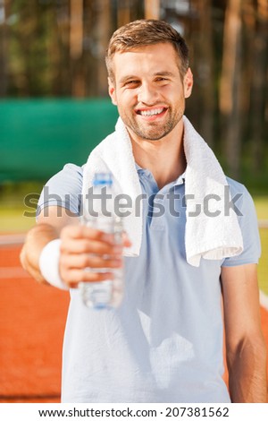 Drink some water! Happy young man in polo shirt and towel on shoulders stretching out bottle with water while standing on tennis court