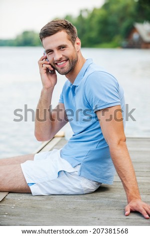Handsome man on the phone. Side view of handsome young man talking on the mobile phone and smiling while sitting at the riverbank