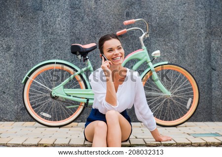 Good talk with friend. Attractive young smiling woman talking on the mobile phone while sitting at the roadside near her vintage bicycle