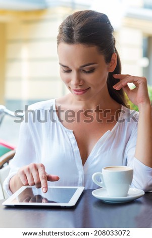 Surfing web in cafe. Beautiful young woman working on digital tablet and smiling while sitting at the sidewalk cafe