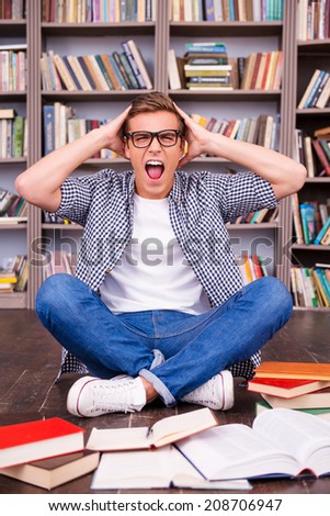 Tomorrow is my final exam! Shocked young man touching his head with hands and shouting while sitting against bookshelf