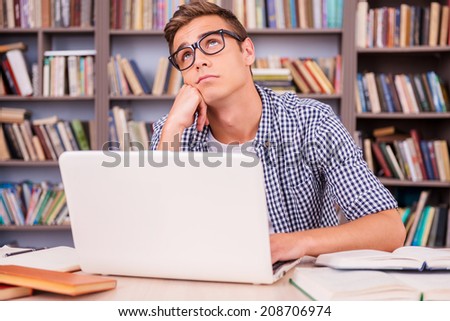 Day dreaming in library. Bored young man leaning his face on hand and looking away while sitting at the desk and in font of bookshelf