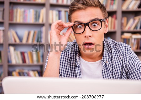 It is unbelievable! Surprised young man looking at laptop and keeping mouth open while sitting against bookshelf