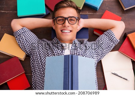 Reading is my hobby! Top view of happy young man holding hands behind head and smiling while lying on the hardwood floor with colorful books laying all around him