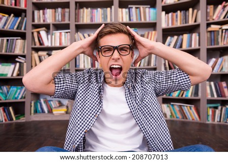 He is not ready to final exams. Shocked young man touching his head with hands and shouting while sitting against bookshelf