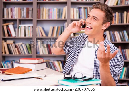 Good talk with friend. Happy young man talking on the mobile phone and gesturing  while sitting at the desk in library