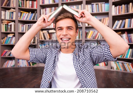 Feeling playful. Cheerful young man carrying book on head and smiling while sitting against bookshelf