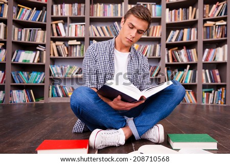 Doing his research in library. Concentrated young man reading book while sitting against bookshelf