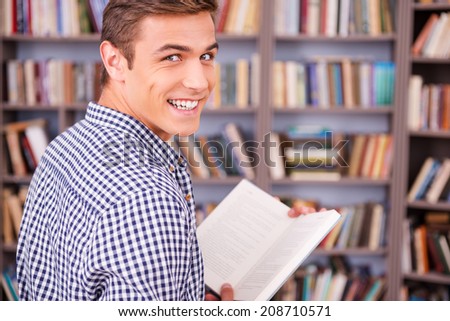 Happy bookworm. Rear view of happy young man holding book and looking over shoulder while standing against bookshelf