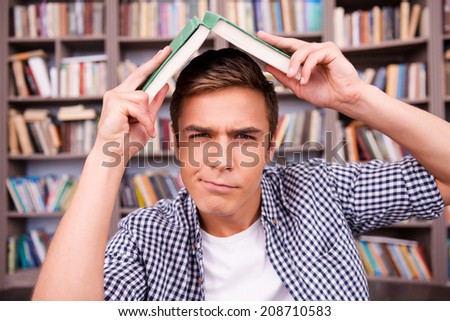 Tired of studying. Frustrated young man carrying book on head and expressing negativity while sitting against bookshelf